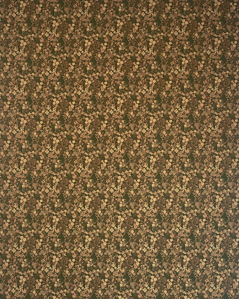The image shows a densely patterned vintage floral wallpaper design by Flat Vernacular. The background is a deep olive green color. The pattern features small clusters of tiny flowers or leaves in muted shades of tan, beige, and brown repeating across the entire surface. The floral elements create a charming, tightly-packed overall texture with an antique, nostalgic aesthetic. The earthy color palette gives a warm, cozy feeling reminiscent of traditional home decor from past eras.