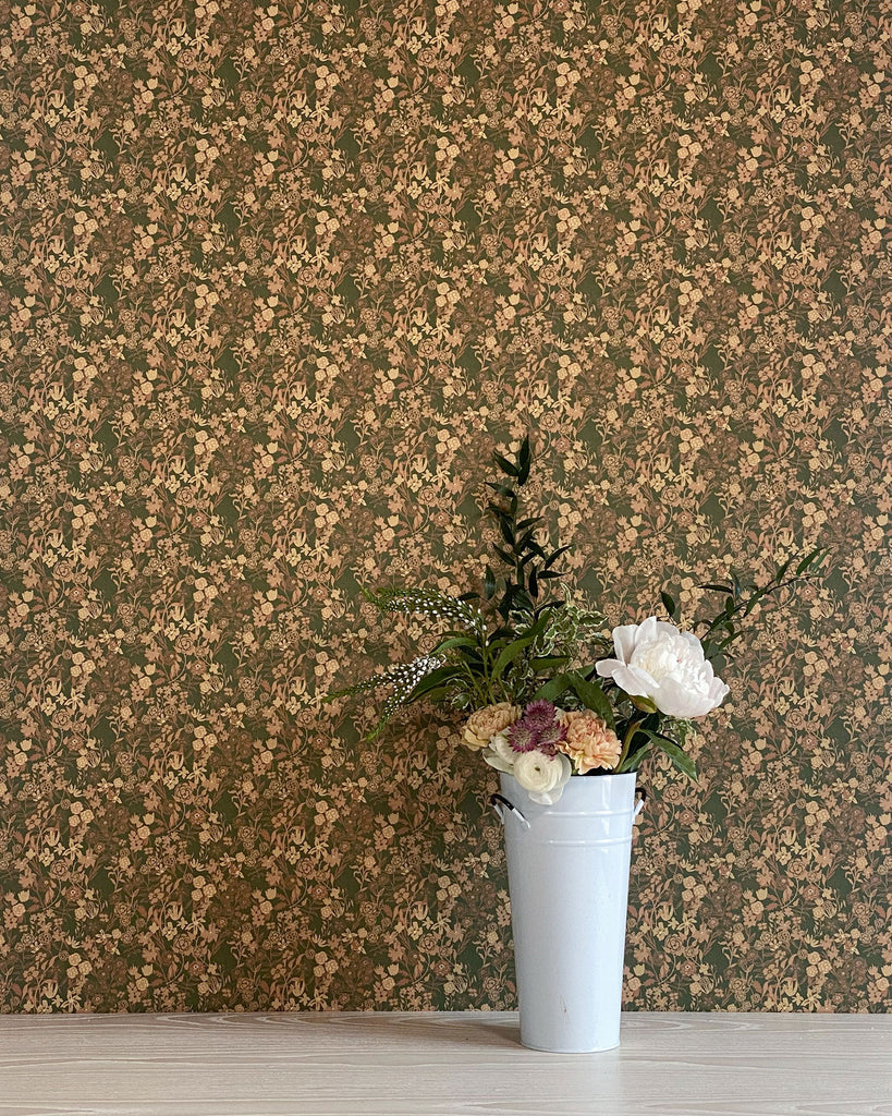 A patterned floral wallpaper design by Flat Vernacular. The background is a deep olive green color. The pattern features small clusters of tiny flowers and leaves in muted shades of tan, beige, and brown repeating across the entire surface. The floral elements create a charming, tightly-packed overall texture with an antique, nostalgic aesthetic.